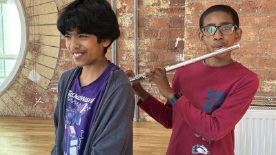 Two young boys, the one on the left is smiling and playing the keyboard, the one on the right is playing the flute, they are in a well lit room with red brick wall